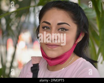 Belle jeune femme Latina avec maquillage moderne pour les yeux et masque rose sous le menton pose pour la caméra et garde la distance sociale pendant la pandémie de corona. Banque D'Images