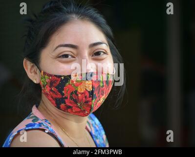 Charmante jeune femme mexicaine positive aux yeux bruns souriants porte un masque facial en tissu non médical coloré pendant la pandémie du coronavirus. Banque D'Images