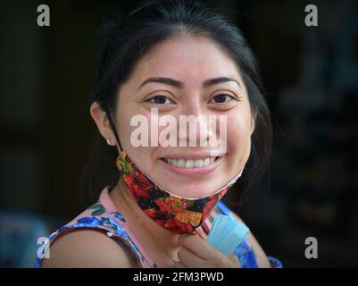 Souriante gaie positive jeune mexicaine Latina femme tire vers le bas son masque de tissu non médical coloré pendant la pandémie mondiale de coronavirus. Banque D'Images