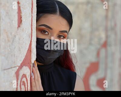 Magnifique modèle mexicain caucasien avec maquillage pour les yeux et tenue noire porte un masque médical noir et regarde autour du coin d'un mur de pierre. Banque D'Images