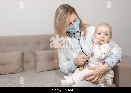 Femme médecin dans le masque bleu faisant un mois de vérification de petite fille tenir un stéthoscope. Une pédiatre féminine examine un enfant mignon, vérifie le cœur et les poumons Banque D'Images