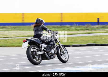 Ulyanovsk, Russie - 10 juin 2017. La jeune fille aux cheveux rouges dans une veste noire et un pantalon de camouflage race sur une moto sur une piste de sport. Flou de mouvement. Banque D'Images