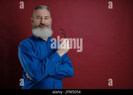 Homme caucasien mûr barbu chemise bleue habillée semble confiant à l'appareil photo avec des lunettes à la main, rouge toile de fond Banque D'Images