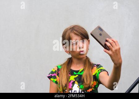 Mise au point automatique. Préteen fille pose et prend un selfie. Adorable petite fille prenant une photo avec un smartphone. Fond gris blanc. Sourire Banque D'Images