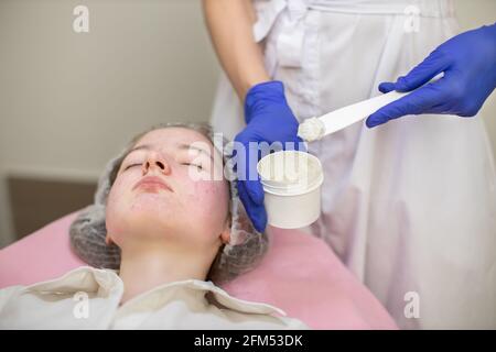 Gros plan d'une jeune adolescente patiente, allongé sur le canapé avec un chapeau jetable à la clinique de dermatologie moderne. Mains de cosmétologue femelle méconnaissable tenant un bocal avec un masque et une spatule Banque D'Images