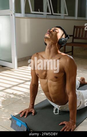 Jeune homme brésilien faisant des cours de yoga en ligne avec le téléphone de tête, dans la position de chien orientée vers le haut, vue de face. Banque D'Images