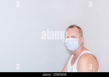 Un homme adulte âgé sous un masque médical dans un T-shirt blanc est assis sur le fond d'un mur gris, regarde vers l'avant. Immunisation de la population, pro Banque D'Images