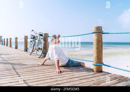 Portrait de l'homme relaxant habillé de vêtements d'été légers et lunettes de soleil assis et profiter du temps et de la vue sur la plage sur le bois pier.négligent vacances à t Banque D'Images