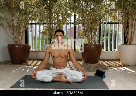 Jeune homme brésilien assis, faisant cours de yoga en ligne en position de méditation. Banque D'Images