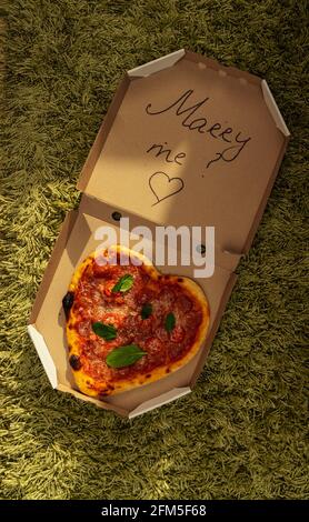 Pizza sous forme de coeur se trouve dans une boîte ouverte avec l'inscription me marier, sur un tapis vert par un jour ensoleillé. Banque D'Images