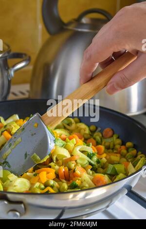 Processus de cuisson faire sauter à la maison. La main de la femme a mélangé des légumes cuits dans une poêle à gaz avec une spatule en silicone et une poignée en bois. Banque D'Images