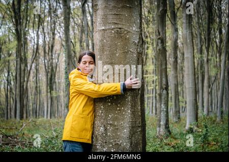 femme en manteau jaune embrassant un arbre dans la forêt avec les yeux fermés. Banque D'Images