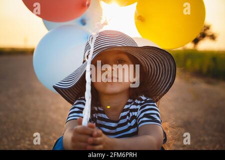 Petite fille malheureuse assise sur la route avec des ballons colorés dedans mains au coucher du soleil Banque D'Images