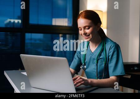 Gros plan de la jeune femme médecin à tête rouge heureuse en bleu vert uniforme médical dactylographiant sur ordinateur portable Banque D'Images