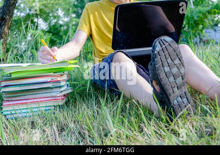 Le gars est un étudiant, un écolier est engagé, fait des devoirs avec un ordinateur portable en ligne dans le jardin sous l'arbre. Banque D'Images