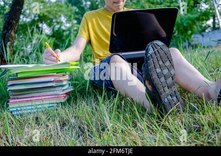 Le gars est un étudiant, un écolier est engagé, fait des devoirs avec un ordinateur portable en ligne dans le jardin sous l'arbre. Banque D'Images