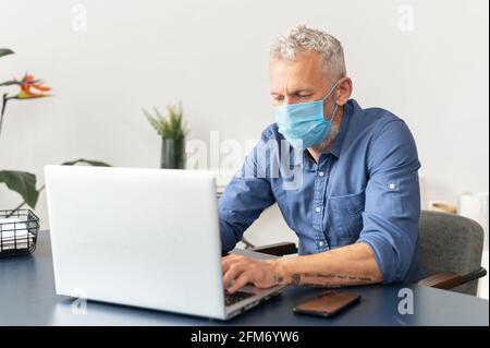 Nouvelle réalité et nouvelles règles pour le personnel de bureau pendant une pandémie. Un employé de sexe masculin mature à cheveux gris portant un masque médical utilise un ordinateur portable au bureau, la prévention de la pandémie et la propagation du virus Banque D'Images