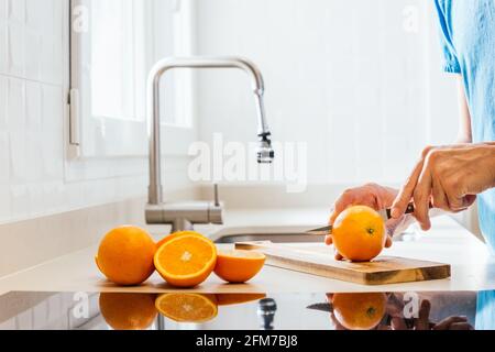 jeune homme en t-shirt bleu coupant des oranges sur un bois planche sur un comptoir de cuisine avec plaque vitrocéramique dans le premier plan et tassement appuyez sur l'arrière-plan avec Banque D'Images