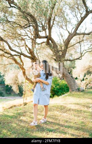 Maman tient doucement son petit fils dans ses bras contre le fond des arbres dans une oliveraie Banque D'Images