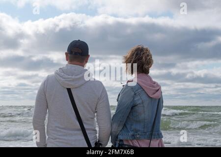 Un couple de jeunes, un homme dans une veste grise et une femme dans une veste bleu denim se tiennent face à la mer par temps venteux sur fond de tempête Banque D'Images