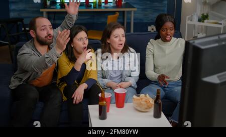 Groupe frustré de personnes multiethniques qui traînaient ensemble tard dans la nuit dans le salon en regardant le football de l'équipe perdre la compétition sportive. Des amis de course mixtes applaudissent à la bière préférée de l'équipe Banque D'Images