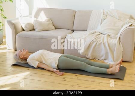 Calme paisible femme sénior pratiquant la Savasana ou le cadavre pose le matin tout en étant allongé sur un tapis de yoga au sol dans le salon à la maison, en longueur. Femme âgée avec les yeux fermés se reposant après l'entraînement Banque D'Images