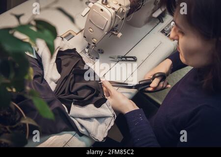 Seamstress rase les vêtements et coupe la couture sur une veste tendance en velours, sur une machine à coudre dans son studio, vue du dessus. Banque D'Images