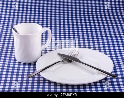 Empy Mug avec cuillère, assiette avec un couteau et fourchette sur la nappe dans une cage. Vue de dessus avec espace de copie Banque D'Images