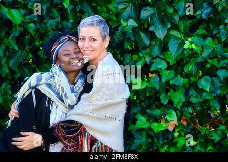 Femme caucasienne mature et jeune femme africaine embrassant dans un jardin avec un fond de feuilles. Banque D'Images