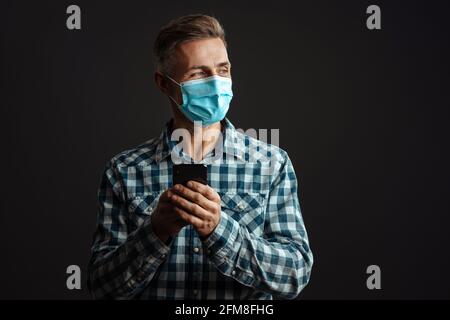 Photo de l'homme optimiste à cheveux gris portant un masque et utilisant téléphone portable isolé sur fond de mur gris Banque D'Images