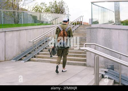 Un jeune homme descend dans la rue avec un skateboard et écoute de la musique avec un casque. Jeunes actifs dans un environnement urbain. Vue arrière Banque D'Images