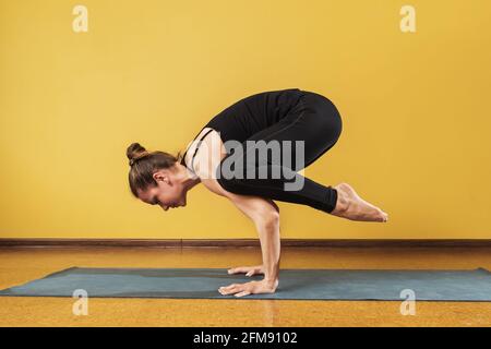 Femme pratiquant le yoga, faisant de l'exercice de bakasana sous forme simplifiée, pose de grue, s'exerçant sur un tapis contre le mur dans le studio. Banque D'Images