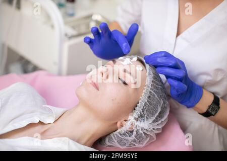 Gros plan vue en angle du visage de la belle jeune femme en chapeau jetable, ayant des procédures de cosmétologie de la peau dans la clinique moderne. Mains du médecin esthéticienne appliquant de la crème sur le visage du client Banque D'Images