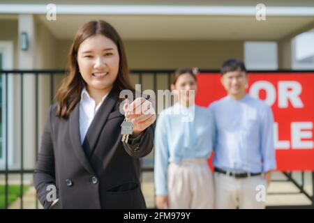 Agent immobilier asiatique ou femme realtor souriant et tenant le dossier rouge avec montrer la clé de maison avec couple asiatique heureux embrassant devant leur maison. Banque D'Images
