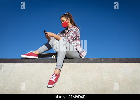 Femme caucasienne portant un masque, assise au mur avec un skateboard et utilisant un smartphone Banque D'Images