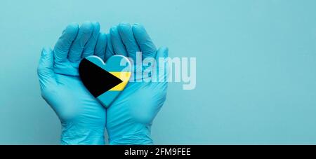 Les médecins mains portant des gants chirurgicaux tenant le coeur du drapeau des Bahamas Banque D'Images