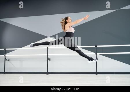 Vue latérale d'une jeune danseuse flexible sautant dans la salle de danse avec des mains courantes de ballet, intérieur high tech. Fille résistante portant des vêtements de sport serrés, qui s'étire et s'exerce dans l'air. Concept sport. Banque D'Images