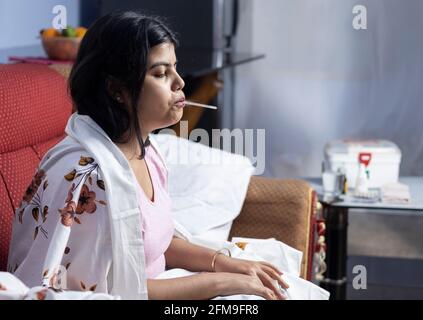 Attention sélective sur une femme indienne assise sur un canapé avec thermomètre dans la bouche pendant la fièvre Banque D'Images