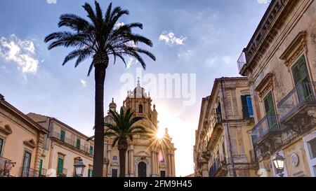 Italie, Sicile, Sicile du Sud-est, angle baroque, Ragusa, Contre-jour le soir, photo HDR, soleil le soir brille de derrière le Duomo di San Giorgio, bâtiments baroques à gauche et à droite le long de la Piazza Duomo, ciel bleu avec des nuages, palmiers ombragés au premier plan Banque D'Images