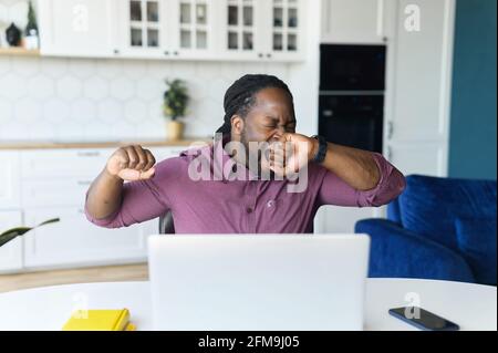 Un travailleur indépendant afro-américain fatigué des années 30 ressent un manque de sommeil et d'énergie, s'assoit au bureau et bâille, bouche couverte de paume, s'étirant les bras sur le lieu de travail. Travail ennuyeux, tôt le matin, travail tardif Banque D'Images