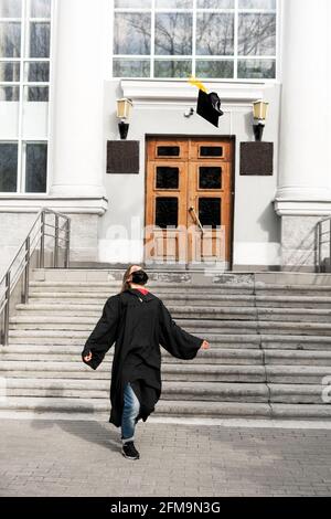 Bonne jeune femme en robe noire de troisième cycle et visage protecteur masque de projection de la casquette de troisième cycle avec pampille jaune sur le fond de l'université ou du collège e Banque D'Images