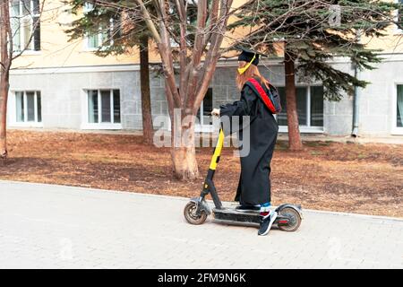 Jeune femme en robe de graduation noire et casquette de graduation noire avec pampille jaune, un scooter électrique après la remise des diplômes à l'université ou coll Banque D'Images