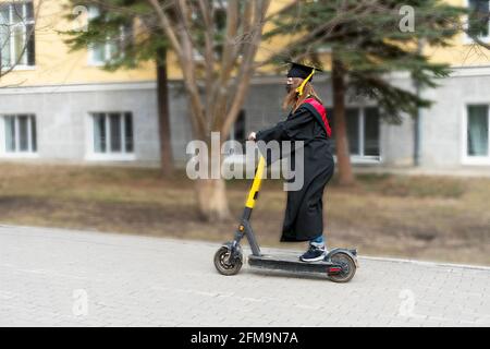 Jeune femme de derrière dans la robe de graduation noire et la casquette de graduation noire avec pampille jaune à cheval un scooter électrique en bas de la rue par l'université Banque D'Images
