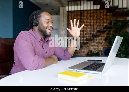 Homme afro-américain heureux avec des serrures cheveux portant un casque sans fil assis au bureau à domicile et saluant les participants de la réunion vidéo, en agitant dans la webcam d'ordinateur portable, tuteur en ligne menant des cours vidéo Banque D'Images