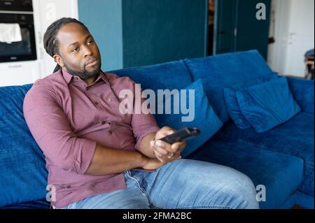 Homme afro-américain ennuyé assis sur le canapé à la maison, en changeant de téléviseur, homme noir endormi et mélancolique tient la télécommande et regarde seul le film sans intérêt à la maison Banque D'Images