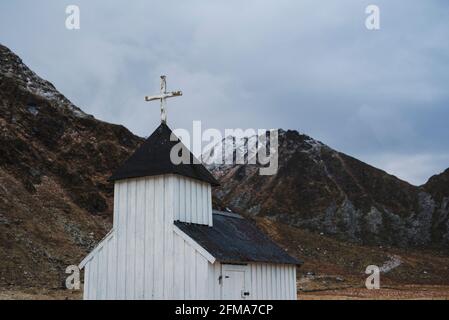 Petite chapelle en bois blanc à Unstad, en face de montagnes légèrement enneigées et de ciel nuageux Banque D'Images