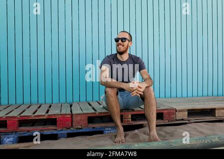 Joyeux jeune barbu portant des lunettes de soleil est souriant à Isla Canela la plage est assise devant un mur en bois bleu vue sur l'extérieur Banque D'Images