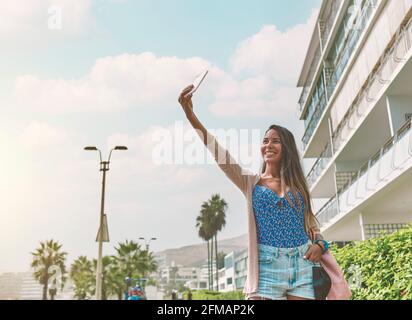 Jeune femme blonde en costume rouge et avec masque protecteur prenant une photo de selfie près du boulevard de luxe et de la plage. Nouveau concept normal Banque D'Images