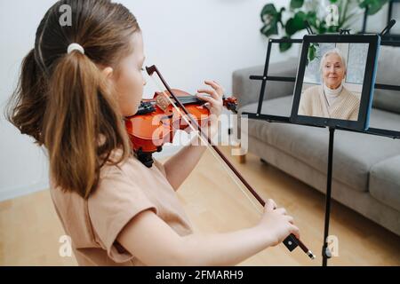 Petite fille avec deux queues de poney apprenant à jouer du violon, montrant son progrès à sa grand-mère dans un chat vidéo. La tablette sur un support de musique. Banque D'Images