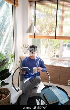 Joyeux jeune homme asiatique dans des vêtements décontractés et des lunettes de lecture réservez tout en vous relaxant dans le salon de sa maison Banque D'Images